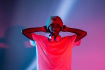 Серьёзная афроамериканка-подросток в наушниках на шее, стоящая в студии с яркими светящимися неоновыми огнями — стоковое фото