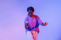 Grave menina adolescente afro-americana com smartphone em fones de ouvido de pé em estúdio com luzes brilhantes de néon — Fotografia de Stock