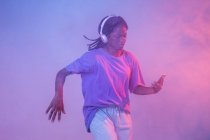 Grave menina adolescente afro-americana com smartphone em fones de ouvido de pé em estúdio com luzes brilhantes de néon e fumaça — Fotografia de Stock
