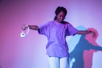 Corpo inteiro de menina adolescente afro-americana com fones de ouvido na mão realizando movimento de dança urbana em estúdio brilhante — Fotografia de Stock