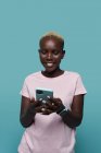 Expresiva hermosa mujer afroamericana con el pelo corto y la navegación de manicura brillante en el teléfono inteligente sobre fondo azul - foto de stock