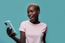 Ausdrucksstarke schöne Afroamerikanerin mit kurzen Haaren und heller Maniküre surft auf Smartphone vor blauem Hintergrund — Stockfoto