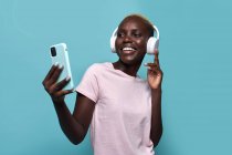 Весёлая африканская американка улыбается, слушая музыку в наушниках на синем фоне — стоковое фото
