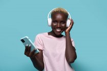 Gai afro-américain femelle dents souriant regarder la caméra tout en écoutant de la musique dans les écouteurs sur fond bleu — Photo de stock