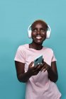 Joyeuse afro-américaine dent féminine souriant tout en écoutant de la musique dans les écouteurs sur fond bleu — Photo de stock