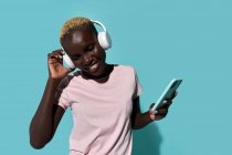 Cheerful African American female toothy smiling while dancing and listening to music in headphones against blue background — Foto stock