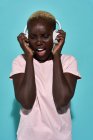 Fröhliche afrikanisch-amerikanische Frau lächelt und singt, während sie Musik mit Kopfhörern vor blauem Hintergrund hört — Stockfoto