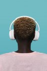 Back view of faceless African American female with short hair listening to music in headphones while standing against blue background — Stock Photo