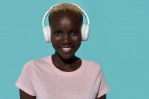 Serene beautiful African American female smiling looking at camera while listening to music in headphones against blue background — Foto stock