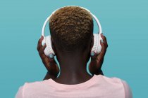 Back view of faceless African American female with short hair listening to music in headphones while standing against blue background — Fotografia de Stock