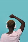 Back view of faceless African American female with raised arm and fist closed listening to music in headphones while standing against blue background — Foto stock
