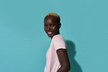 Side view of serene cute beautiful African American female smiling looking at camera standing against blue background in a studio - foto de stock
