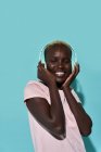 Gai afro-américain femelle dents souriant avec les yeux fermés en écoutant de la musique dans les écouteurs sur fond bleu — Photo de stock