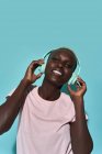 Cheerful African American female toothy smiling looking at camera listening to music in headphones against blue background — Stock Photo