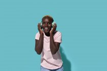 Cheerful African American female toothy smiling looking at camera listening to music in headphones against blue background — Fotografia de Stock