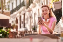 Весела жінка з бездротовими навушниками на шиї серфінг інтернет на мобільному телефоні сидячи за столом у кафе на вулиці в божевільні — стокове фото