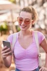 Счастливая женщина в солнечных очках смотрит мобильный телефон, стоя в уличной столовой в Испании — стоковое фото
