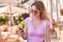 Happy female in sunglasses watching mobile phone while standing in street cafeteria in Spain - foto de stock
