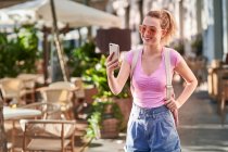 Щаслива жінка у сонцезахисних окулярах дивиться на мобільний телефон, стоячи у вуличній їдальні на шпині. — стокове фото