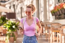 Щаслива жінка у сонцезахисних окулярах дивиться на мобільний телефон, стоячи у вуличній їдальні на шпині. — стокове фото