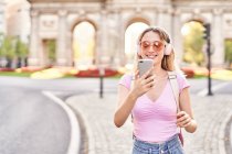 Femme gaie en tenue décontractée et lunettes de soleil prenant autoportrait tout en écoutant de la musique sur la rue madrid — Photo de stock