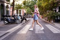 Vista lateral cuerpo completo de mujer joven caminando por el paso peatonal y comprobando ruta en teléfono móvil en Madrid - foto de stock