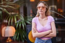 Concerned female in sunglasses wearing casual clothes standing with crossed arms against glass wall in Madrid street — Stock Photo