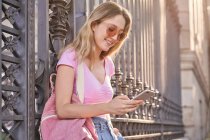 Вид збоку на позитивну жінку з рюкзаком, який яскраво посміхається, прокручуючи мобільний телефон, що спирається на металевий паркан — стокове фото