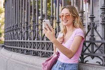 Side view of positive female with backpack smiling brightly while scrolling mobile phone leaning on metal fence — Stock Photo