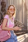 Side view of positive female with backpack smiling brightly while scrolling mobile phone leaning on metal fence — Stock Photo