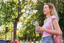 Seitenansicht einer fröhlichen jungen Frau mit Rucksack und Wasserflasche, die im grünen Park an einem sonnigen Tag in Madrid steht — Stockfoto