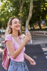 Vista lateral de jovem alegre com mochila e garrafa de água em pé no parque verde em dia ensolarado em Madrid — Fotografia de Stock