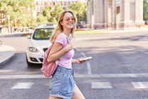 Вид сбоку молодой женщины, идущей по пешеходному переходу и проверяющей маршрут по мобильному телефону в Мадриде — стоковое фото