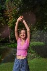 Mujer alegre con los brazos levantados de pie en salpicaduras en el parque soleado - foto de stock