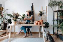 Молодая женщина-садовод в соломенной шляпе создает букет на столе с разнообразными инструментами дома — стоковое фото