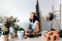 Young female horticulturist in straw hat sitting near flowers on table with assorted tools at home - foto de stock