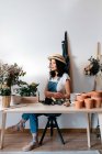Jeune horticultrice en chapeau de paille assise près des fleurs sur la table avec des outils variés à la maison — Photo de stock