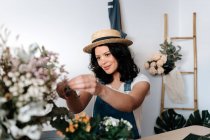 Jeune horticultrice en chapeau de paille créant un bouquet sur la table avec des outils variés à la maison — Photo de stock