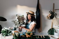 Молодая женщина-садовод в соломенной шляпе создает букет на столе с разнообразными инструментами дома — стоковое фото