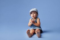 Criança descalça alegre encantadora em vestido de ganga e chapéu com cabelo encaracolado olhando para longe enquanto joga no fundo azul — Fotografia de Stock