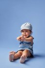 Очаровательный веселый босиком ребенок в джинсовом платье и шляпе с вьющимися волосами, глядя в сторону, играя на синем фоне — стоковое фото