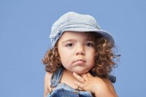 Adorable niño molesto con ojos marrones usando ropa casual y sombrero contra fondo azul mirando hacia otro lado - foto de stock