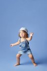 Criança descalça encantadora em vestido de ganga e chapéu com cabelo encaracolado olhando para longe enquanto dança no fundo azul — Fotografia de Stock