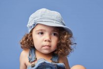 Чарівна засмучена маленька дитина з коричневими очима в повсякденному одязі і капелюсі на синьому фоні, дивлячись геть — стокове фото