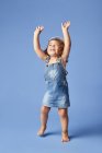 Очаровательный босоногий ребенок в джинсовом платье и шляпе с вьющимися волосами, смотрящий вверх с поднятыми руками, танцуя на синем фоне — стоковое фото