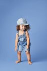 Charmantes barfüßiges Kind in Jeanskleid und Hut mit lockigem Haar, das beim Tanzen auf blauem Hintergrund wegschaut — Stockfoto