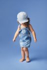 Очаровательный босоногий ребенок в джинсовом платье и шляпе с вьющимися волосами, смотрящий в сторону, танцуя на синем фоне — стоковое фото