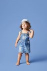 Очаровательный босоногий ребенок в джинсовом платье и шляпе с вьющимися волосами, смотрящий в сторону, танцуя на синем фоне — стоковое фото