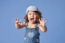 Charmantes barfüßiges Kind in Jeanskleid und Hut mit lockigem Haar, das vor blauem Hintergrund in die Kamera schaut und Gesichter macht — Stockfoto