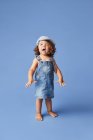 Charmant fröhliches Barfußkind in Jeanskleid und Hut mit lockigem Haar, das in die Kamera schaut, während es auf blauem Hintergrund tanzt — Stockfoto